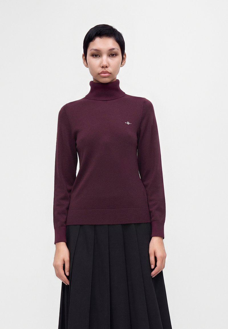 Femme portant un pull à col roulé bordeaux avec un petit emblème sur la poitrine et une jupe noire plissée, debout devant un fond clair uni.