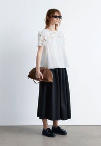 White blouse with lace detailing, black pleated maxi skirt, brown suede handbag, and black patent loafers. Model wearing black sunglasses.