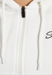 Witte zip-up hoodie van een textuurstof, met een zwarte metalen rits en witte aantrekkoorden. Logo geborduurd aan de linkerkant.
