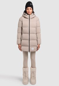 Jachetă puffer bej cu fermoar frontal și glugă, purtată împreună cu colanți bej și cizme albe masive. Căciulă tricotată neagră pe cap.