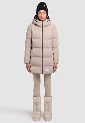Beige pufferjack met een voorzijde rits en capuchon, gecombineerd met beige legging en chunky witte laarzen. Zwarte gebreide beanie op het hoofd.