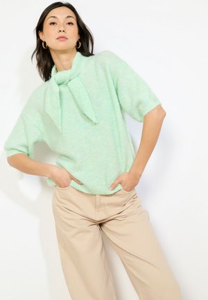 Žena oblečená v krátkom ružovozelenom svetri s kravatovým golierom a béžových vysokých nohaviciach, stojaca s rukami v kapsách.