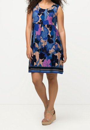 Vrouw draagt mouwloze knielange jurk met blauw, paars en beige bloemenpatroon, gecombineerd met beige sleehak sandalen.