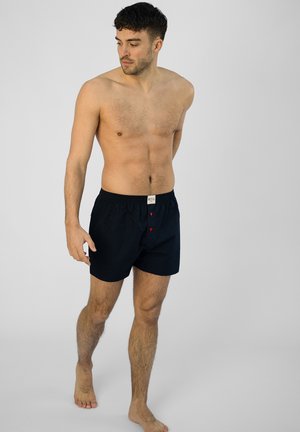 Marineblaue Baumwollboxershorts mit einem elastischen Bund und zwei roten Knöpfen vorne. Der Stoff hat eine glatte Textur.