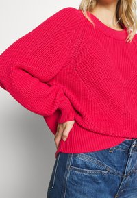 Femme portant un pull en maille rose vif texturé à manches larges et un jean en denim bleu, main posée sur la hanche.
