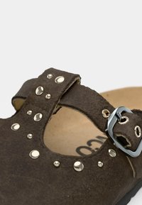 Καφέ δερμάτινο σανδάλι με φαρδιά λουρί που διαθέτει διακοσμητικά μεταλλικά τρουκ και μεταλλικό πόμολο. Μαλακό πέλμα ορατό στο εσωτερικό.