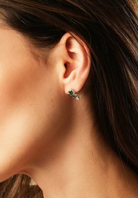 Kvindes ører med mørkebrunt hår iført sølvringe, der har to grønne og en klar ædelsten.