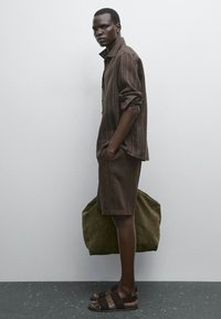 Chemise à rayures brunes, short brun, sandales noires ; portant un sac vert olive. Le modèle se tient contre un mur gris clair sur un sol sombre.