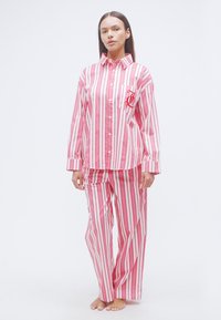 Σετ πιτζάμες με ρίγες σε ροζ και λευκό, που περιλαμβάνει πουκάμισο με κουμπιά και τσέπη με κόκκινο λογότυπο, καθώς και παντελόνι με ελαστική μέση που ταιριάζει.