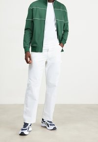 Giacca verde con zip e dettagli bianchi, indossata sopra una maglietta bianca, abbinata a pantaloni bianchi a vita alta e scarpe da ginnastica blu e bianche.