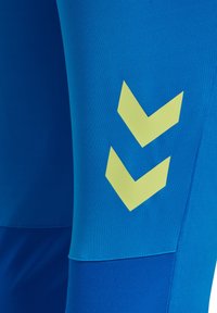 Niebieskie legginsy sportowe o gładkiej teksturze z kontrastowym, żółtym logo w kształcie chevrona na udzie. Bezszwowy krój w pasie.
