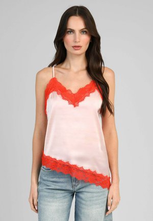 Femme aux longs cheveux foncés portant un caraco en satin rose clair avec une bordure en dentelle rouge et un jean bleu clair, debout devant un fond gris.