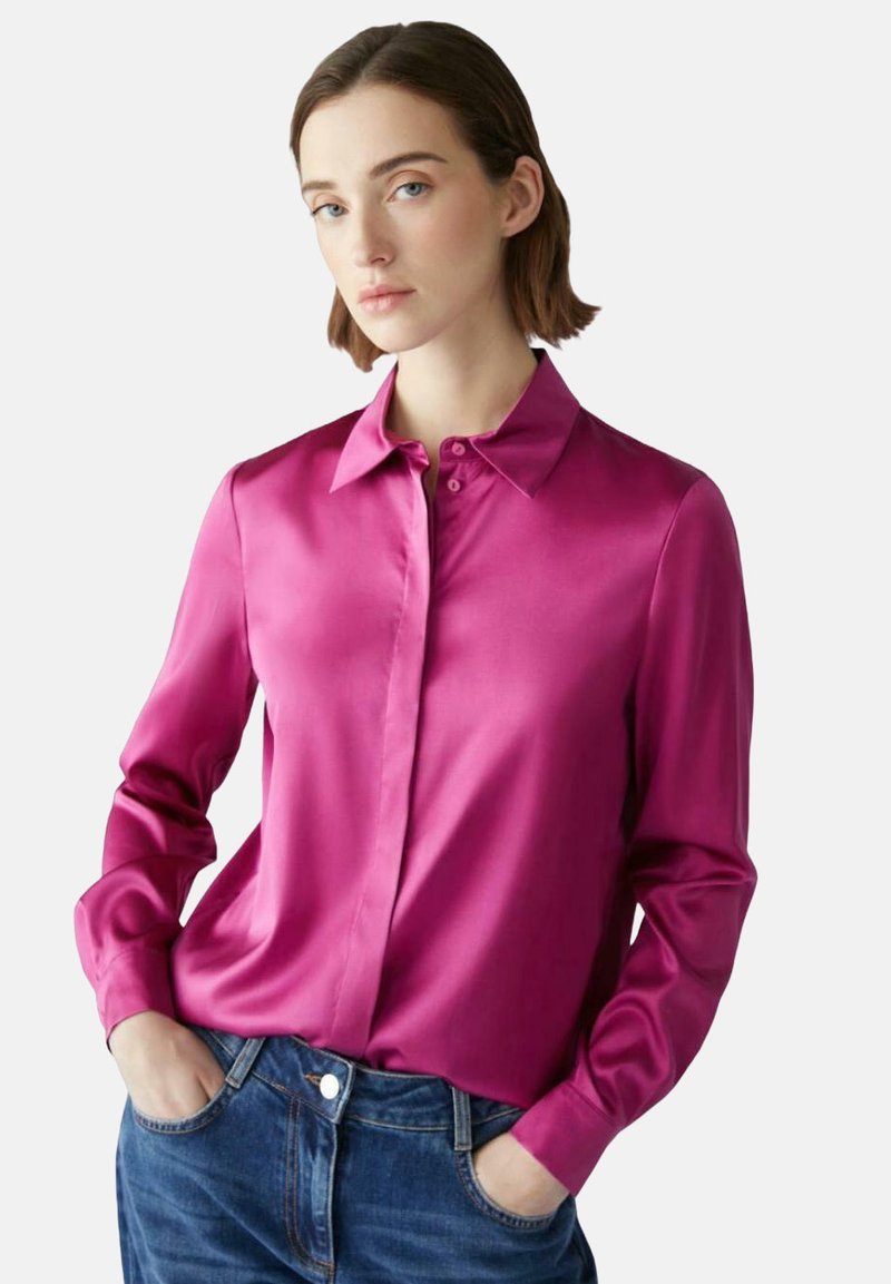 Fuchsia satijnen blouse met een puntige kraag, lange mouwen en knoopsluiting aan de voorkant. Gecombineerd met blauwe jeans, die een relaxed fit laten zien.