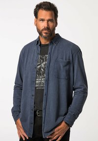 Camicia blu navy con una morbida texture, maniche lunghe e taschino sul petto, sovrapposta a una t-shirt nera con grafica. Cintura nera e pantaloni scuri.