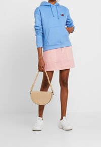 Felpa azzurra chiara con tasca frontale, abbinata a una gonna di jeans rosa. Tiene una borsa a mezzaluna beige e testurizzata con manico intrecciato. Sneakers bianche.