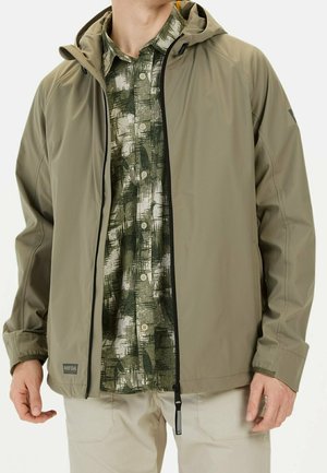 Homme portant une veste beige à capuche par-dessus une chemise verte à motifs boutonnée et un pantalon beige clair, debout avec les mains détendues le long du corps.