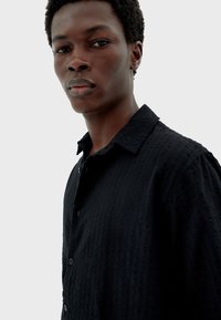 Schwarzes Hemd mit Knöpfen und strukturiertem Muster, mit Kragen und kurzen Ärmeln, präsentiert an einem Modell vor einem schlichten Hintergrund.