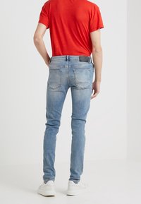 Jeans de denim azul claro con un corte ajustado, diseño de cinco bolsillos, presentados con un parche de cuero negro en la cinturilla y textura desgastada.
