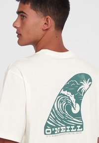 Cream-colored t-shirt featuring a green wave graphic with palm trees and sun. Relaxed-fit with short sleeves and a crew neckline.