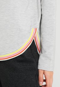 Top de manga larga gris claro con dobladillo curvado, acentuado por un ribete de rayas multicolores en rosa, amarillo y naranja en los lados.