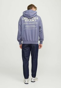 Hoodie in lichtpaars met bedrukte tekst op de achterkant, gecombineerd met donker denim broek en lichte sneakers. Zacht textiel.