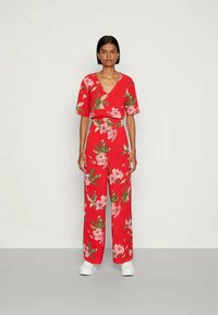 Macaquinho floral vermelho com mangas curtas, decote em v e cintura cinturada. Apresenta flores cor-de-rosa e folhas verdes sobre um fundo brilhante.