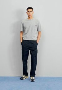 Grå kortærmet t-shirt med lomme, parret med mørke bukser og sorte/gule sneakers. Stående foran en ensfarvet grå baggrund.