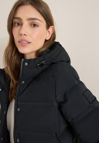 Doudoune noire avec capuche, dotée d'une fermeture éclair et de boutons-pression. Texture lisse avec un patch logo sur la manche.