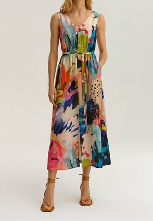 Femme portant une robe sans manches à imprimé abstrait coloré avec une ceinture à la taille et des sandales à lanières beiges sur un fond uni.