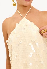 Femme portant un top halter crème recouvert de grands sequins ronds, tenant le tissu, avec une boucle d'oreille dorée en forme de coquillage et une bague dorée visibles.