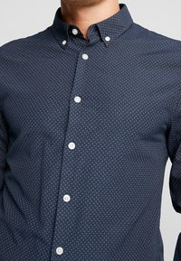 Man som bär en marinblå skjorta med små vita diamantmönster och vita knappar, kragen knäppt, utan synlig bakgrund.