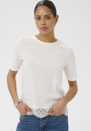 Femme aux cheveux foncés portant un haut blanc à manches courtes avec un ourlet en dentelle, un jean bleu, un bracelet argenté et de petites boucles d'oreilles anneaux, debout les mains dans les poches.