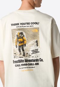 Jasna kremowa bawełniana koszulka z grafiką przedstawiającą osobę w żółtej kurtce w zaśnieżonym otoczeniu, z wyraźnym tekstem oraz logo The North Face.