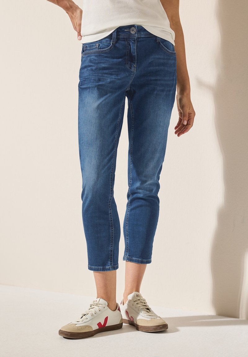 Personne portant un jean skinny bleu court avec des détails délavés et des baskets blanches avec des accents rouges, debout contre un mur clair uni.