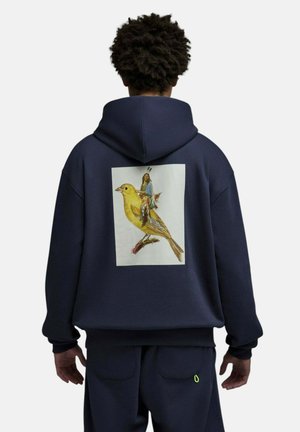 Personne portant un sweat à capuche bleu marine et un pantalon, le dos du sweat présente un grand graphisme d’un oiseau jaune avec une petite figure humaine debout dessus.