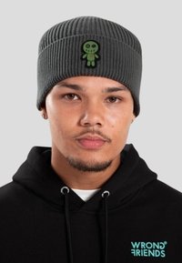 Mladý muž v šedé pletené čepici s zeleným vyšitým štítkem panenky a černé mikině s logem "Wrong Friends".