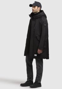 Schwarzer Parka mit Kapuze, ausgestattet mit verstellbaren Kordeln, Druckknöpfen und zwei Seitentaschen. Getragen mit schwarzen Jeans und Schuhen.