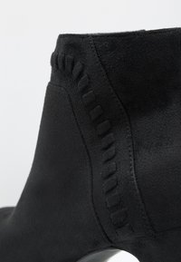 Gros plan sur une botte cheville en daim noir montrant un détail décoratif de couture le long du côté et un talon arrondi lisse.