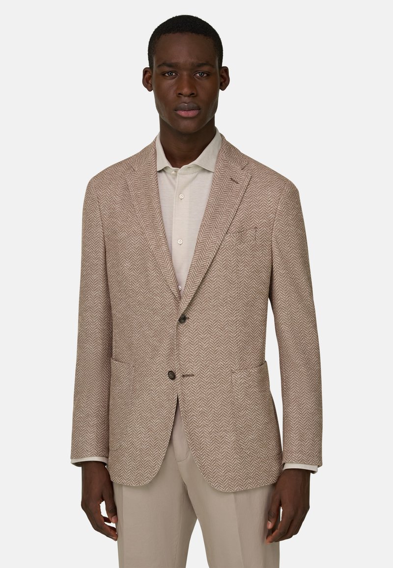 Mann trägt einen beigen Herringbone-Blazer über einem hellcremefarbenen Hemd mit Knöpfen und beigen Hosen, steht vor einem schlichten Hintergrund.