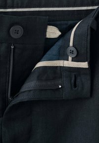 Ceinture de pantalon noire avec fermeture éclair dézippée, deux boutons noirs, bordure beige et boucle de tissu beige visible sur la doublure intérieure.
