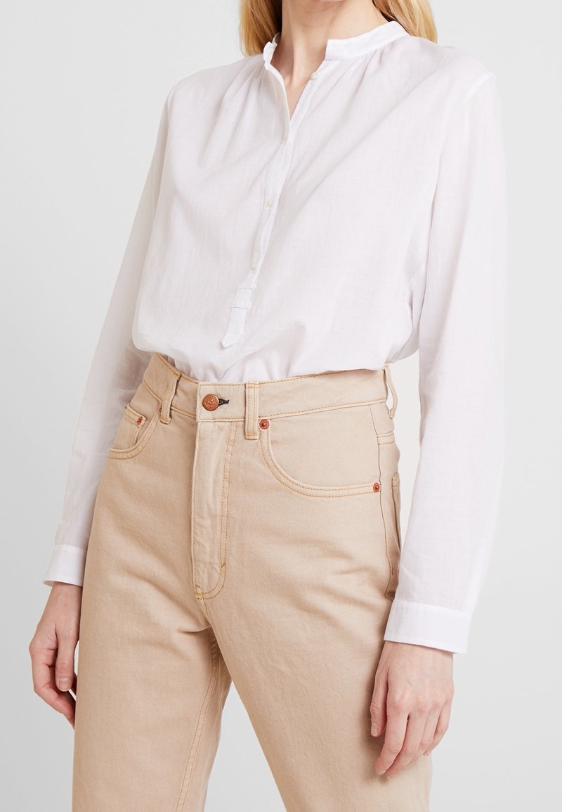 Femme portant une chemise blanche à manches longues boutonnée rentrée dans un jean beige taille haute sur un fond uni.
