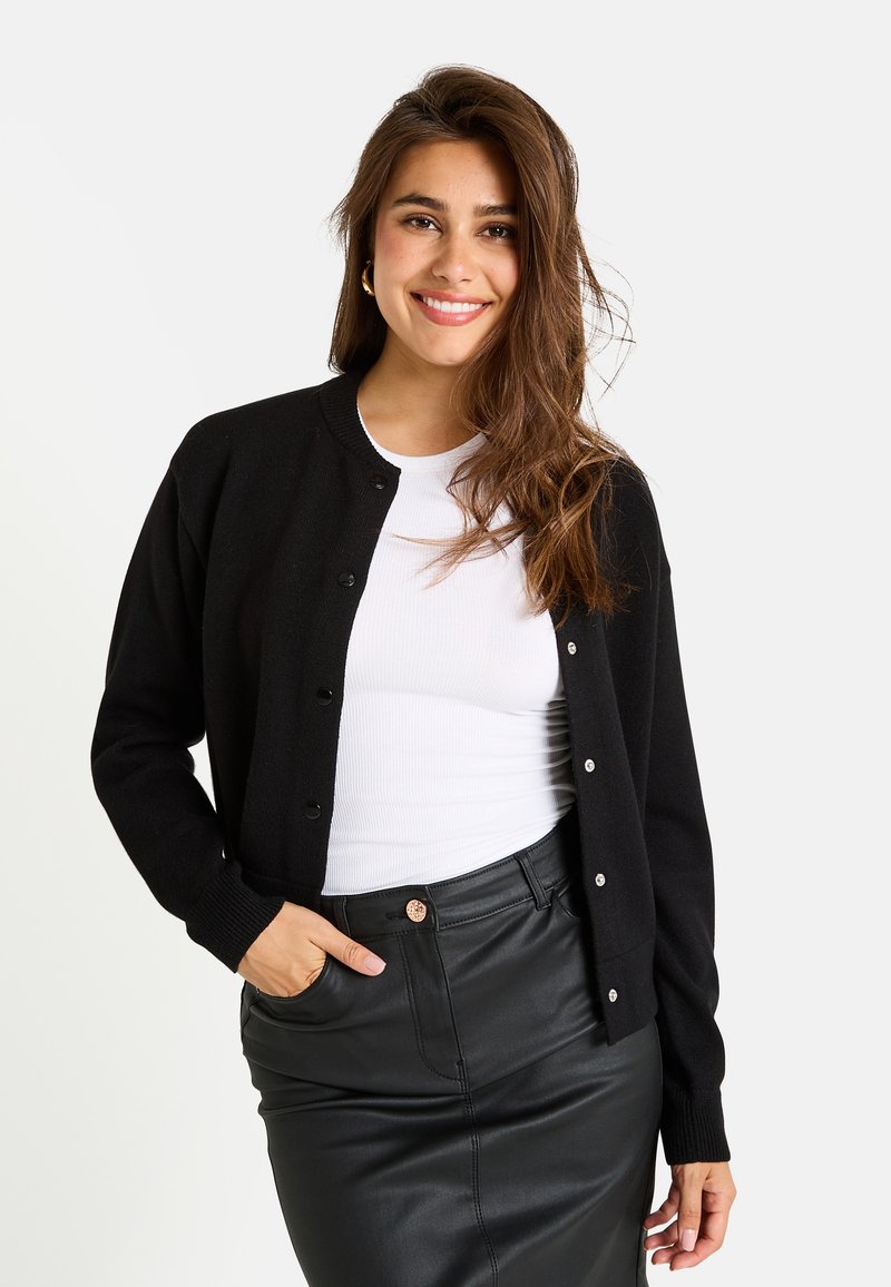 Cardigan en maille noire avec un col ronde, des boutons sur le devant et des poignets côtelés. Associé à un haut blanc et une jupe en cuir noir.