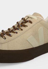 Baskets décontractées beige avec semelle marron texturée, lacets marron et bleu tachetés, et logo crème "V" sur le côté.