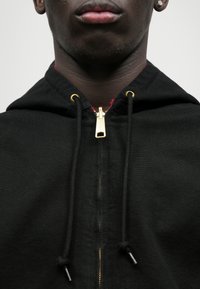 Črna zip-up kapuca s zlatim zadrgo, nastavljivimi vrvicami in vidno rdečo podlogo. Izdelana iz mešanice bombaža.