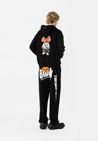 Conjunto de sudadera con capucha negra y pantalones que presenta un gráfico de un oso con ropa deportiva. Los pantalones tienen texto blanco y una correa decorativa en el lado.