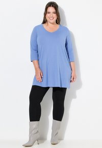 Helles blaues Tunika-Oberteil mit V-Ausschnitt und Dreiviertelärmeln, kombiniert mit schwarzen Leggings und grauen spitzen Stiefeletten.