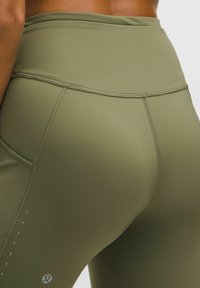 Primo piano di una persona che indossa leggings verde oliva a vita alta con cuciture visibili e un piccolo logo sulla coscia.