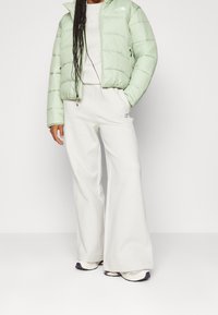 Jasnozielona kurtka puchowa z zamkiem z przodu i bocznymi kieszeniami, w połączeniu z białymi spodniami dresowymi o szerokich nogawkach. Sneakersy z fioletową podeszwą dopełniają całość.