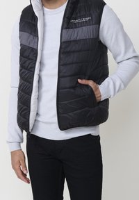 Homme portant un gilet matelassé noir et gris sur une chemise blanche texturée à manches longues, une main dans la poche du gilet.