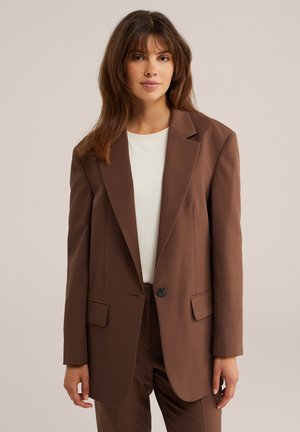 Donna con capelli castani fino alle spalle che indossa un blazer marrone e pantaloni coordinati sopra una camicia bianca, in piedi davanti a uno sfondo neutro.